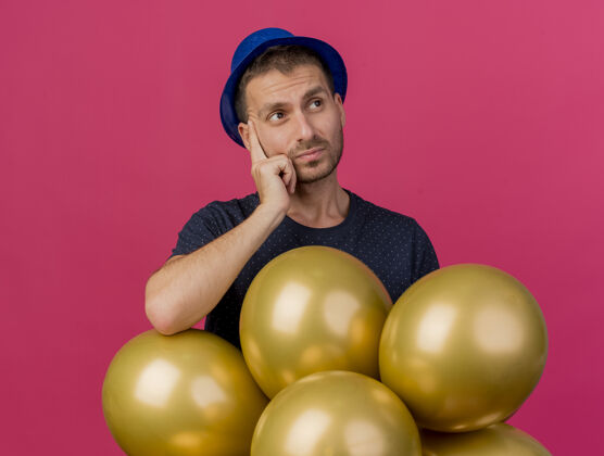 站迷茫的帅哥戴着蓝色的派对帽拿着氦气球看着一面隔离在粉色墙上的复制空间人氦脸