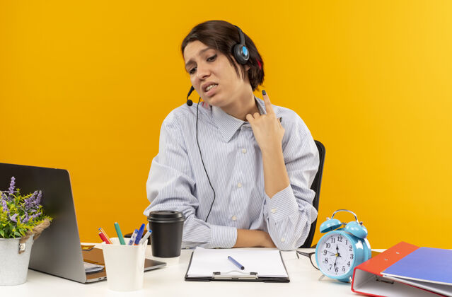 举起年轻的电话中心女孩戴着耳机坐在办公桌旁 举着工作工具 孤立地看着橙色的背景电话工作居中