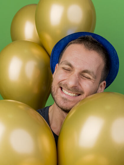 微笑笑容可掬的帅哥头戴蓝色派对帽 站着氦气球 隔离在绿色墙壁上 留有临摹空间表情氦人