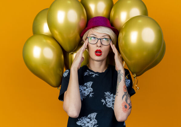 人疲惫的金发派对女郎戴着派对帽戴着眼镜站在气球前 手举在头上孤立地看着橙色的墙上金发站帽子