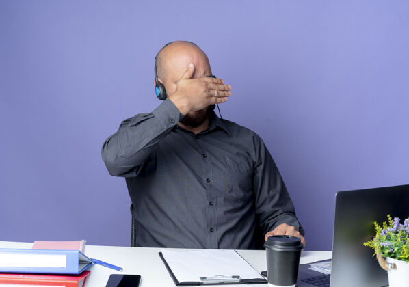 年轻年轻秃顶的呼叫中心男子戴着耳机坐在办公桌旁 工作工具盖在脸上 手被隔离在紫色背景上秃头脸坐着