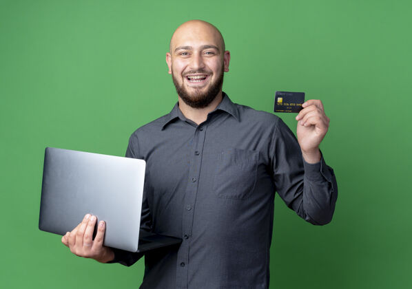 中心快乐的年轻秃头呼叫中心男子手持笔记本电脑和信用卡隔离在绿色控股男人绿色