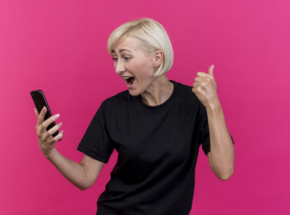 脸快乐的中年金发斯拉夫妇女抱着手机 看着手机显示拇指向上孤立的粉红色墙上衣服站着人
