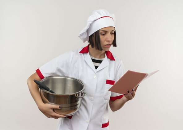 烹饪年轻的女厨师穿着厨师制服拿着锅子和便笺簿看着隔离在白色背景上的便笺簿女垫锅