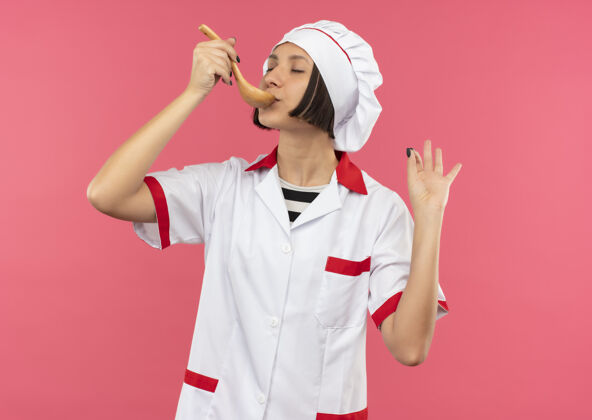 烹饪身着厨师制服的年轻女厨师正在用勺子吃饭 在粉色背景上闭着眼睛 做着“ok”的手势厨师关门眼睛
