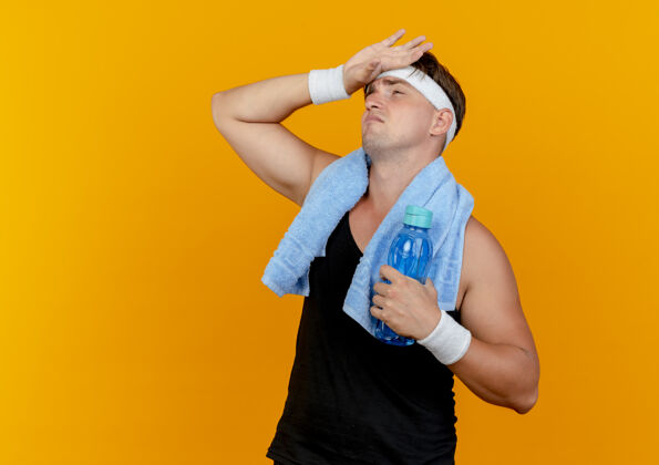 戴着疲惫的年轻帅气的运动型男人戴着头带和手镯 脖子上围着毛巾 手放在额头上 手拿着水瓶 在橙色背景上隔离 留有复制空间头带围着抱着