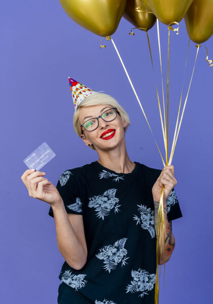 人微笑着的金发派对女郎戴着眼镜 戴着生日帽 手里拿着气球和信用卡 看着前面孤立的紫色墙壁聚会人立场
