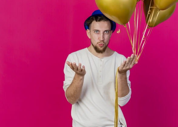 年轻自信的年轻帅哥斯拉夫党的家伙戴着党的帽子拿着气球看前面显示空手做亲吻手势孤立在粉红色的墙上复制空间斯拉夫姿势聚会
