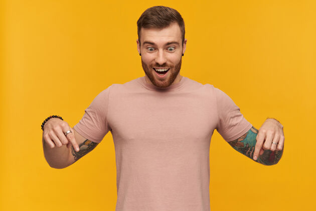 站一个快乐英俊的年轻人 穿着粉色T恤 留着胡子 手上有纹身 用两个手指指着黄色的墙上墙指向手