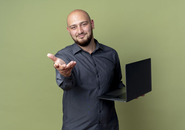 伸展高兴的年轻秃顶呼叫中心男子手持笔记本电脑 伸出手对着相机隔离在橄榄绿的背景与复印空间抱着请橄榄