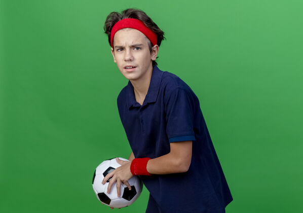 球皱眉的年轻帅气的运动男孩戴着头带和护腕 戴着牙套 拿着足球 看着前面隔离在绿色墙上的复制空间姿势足球皱眉