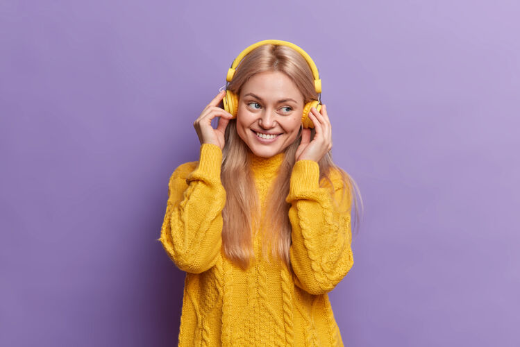 应用程序积极向上的千禧一代女孩戴着耳机享受着悦耳的音乐心情愉快地微笑着穿着黄色套头衫快乐室内耳机
