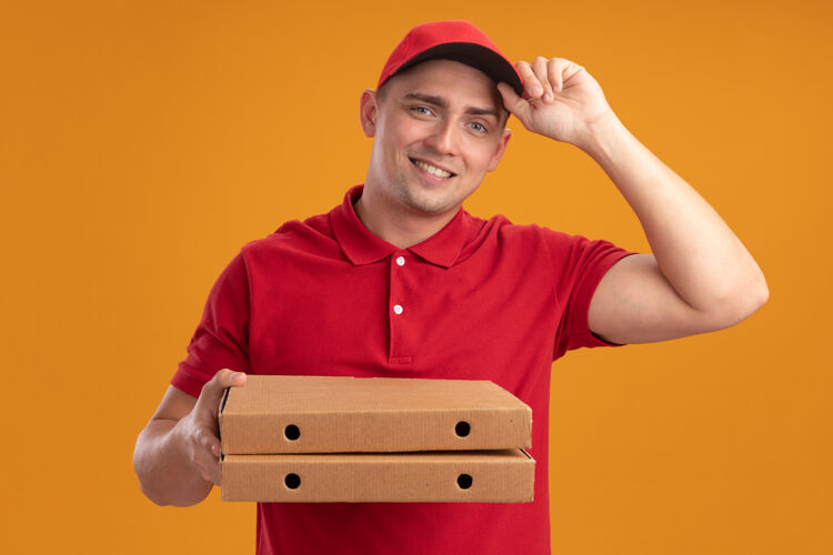 男人微笑着的年轻送货员穿着制服 戴着帽子 拿着隔离在橙色墙上的比萨饼盒送货微笑年轻人