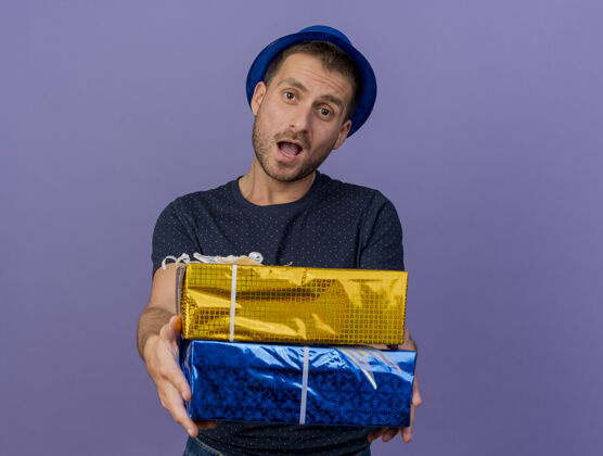 持有印象深刻的英俊白人男子戴蓝色帽子举行礼品盒孤立的紫色背景与复制空间印象礼品盒子