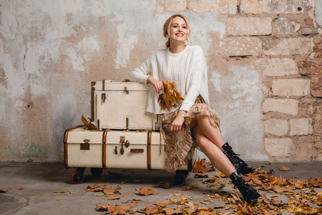 树叶迷人的微笑 穿着白色针织毛衣的时尚金发女人坐在手提箱上的肖像在街上的老式墙上手提箱休闲城市