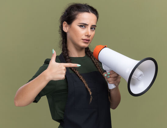 信心自信的年轻女理发师拿着制服 指着隔离在橄榄绿墙上的扬声器制服举行年轻