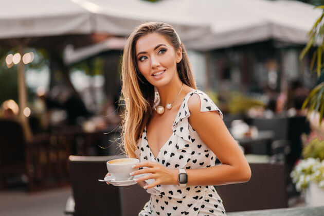 餐厅美丽的微笑的女人穿着时尚的白色印花连衣裙坐在街上的咖啡馆里喝着一杯卡布奇诺可爱桌子夏天