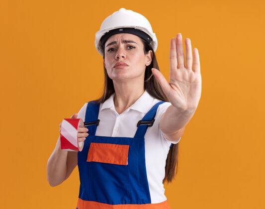 年轻穿着制服的严格的年轻建筑妇女拿着管道胶带 在橙色的墙上显示停止手势举行女人显示