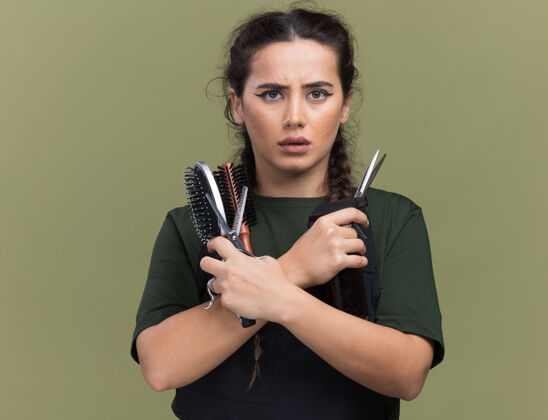 工具自信的年轻女理发师穿着制服拿着和交叉理发工具隔离在橄榄绿的墙上橄榄理发师拿着