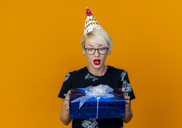 空间令人印象深刻的年轻金发派对女孩戴着眼镜和生日帽举行 看着礼品盒孤立的橙色背景与复制空间礼物金发眼镜