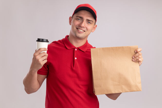 年轻人带着微笑的年轻送货员 穿着制服 戴着帽子 手里拿着一个纸制食品包 白墙上隔着一杯咖啡男人咖啡帽子