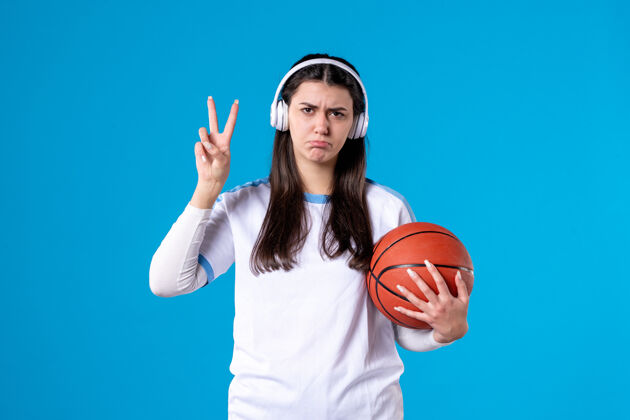 人正面图：戴着耳机拿着篮球的年轻女性站在蓝色的墙上青年肖像微笑