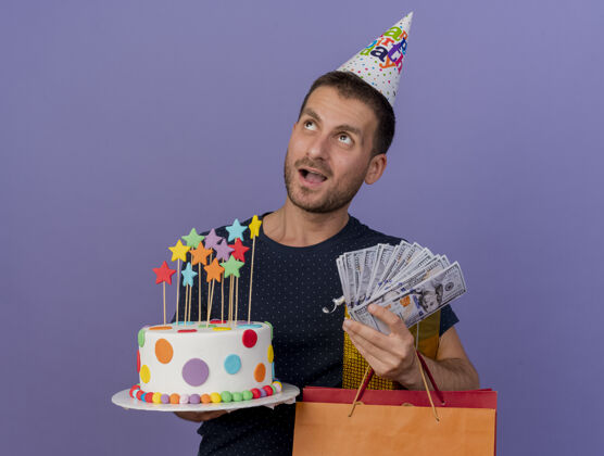 复制兴奋帅气的白种人戴着生日帽拿着生日蛋糕纸购物袋礼盒和钱看着边上孤立的紫色背景和复制空间蛋糕空间帽子