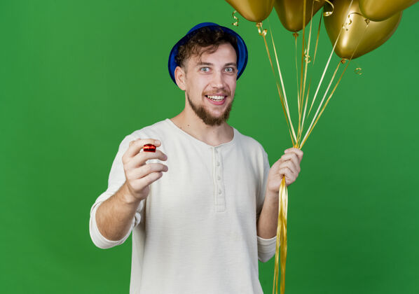 小伙子面带微笑的年轻英俊的斯拉夫党人戴着党的帽子拿着气球和党吹在绿色背景与复制空间隔离相机帽子绿色吹风机