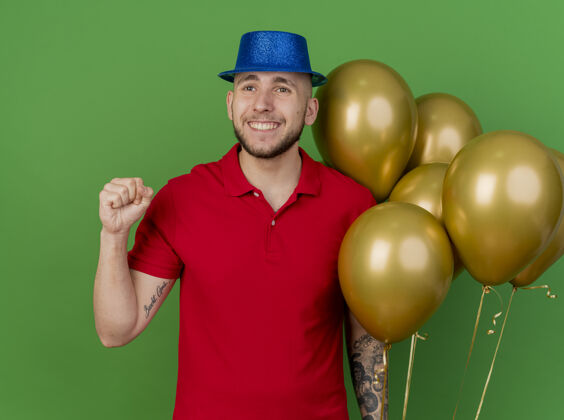 气球面带微笑的年轻英俊的斯拉夫党人戴着党的帽子拿着气球看着一边做是的手势孤立的绿色背景绿色年轻派对