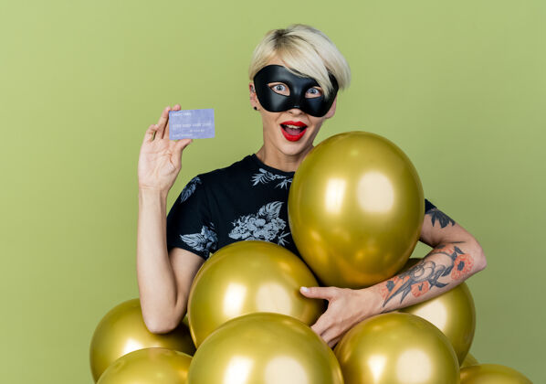 派对令人印象深刻的年轻金发派对女孩戴着化装面具站在气球后面显示信用卡看着橄榄绿背景上孤立的相机印象面具化装舞会