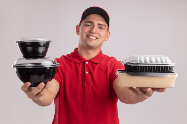 穿微笑着的年轻送货员穿着制服 戴着帽子 举着食品容器 对着隔离在白墙上的摄像机制服男人食物