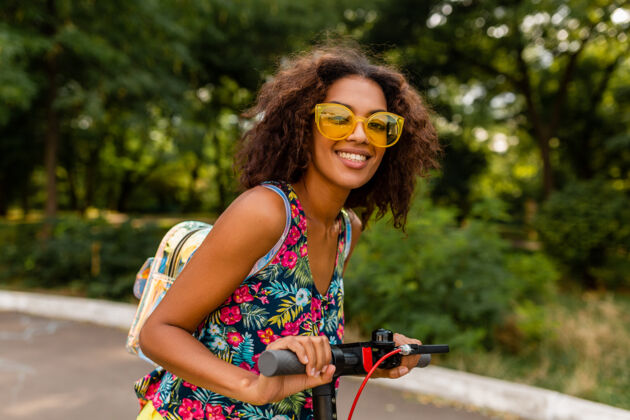 欢呼年轻时尚的黑人女子在公园里玩得很开心骑着电动脚踏车在夏天的时尚风格 五颜六色的时髦服装 穿着背包和黄色太阳镜道路女性积极