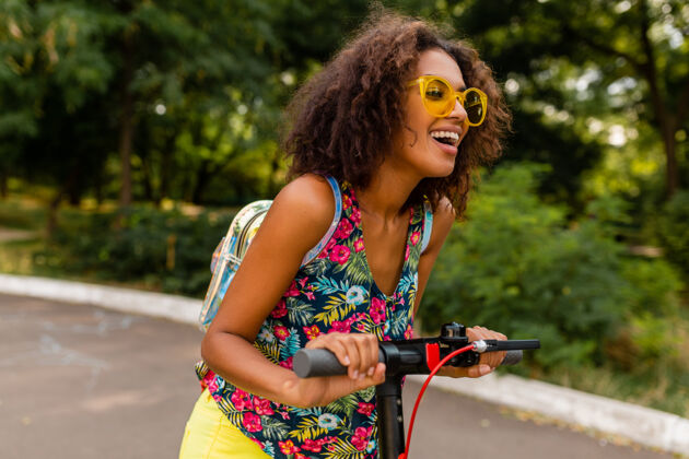 女孩年轻时尚的黑人女子在公园里玩得很开心骑着电动脚踏车在夏天的时尚风格 五颜六色的时髦服装 穿着背包和黄色太阳镜环保兴奋车辆