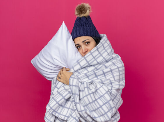 深红色虚弱的年轻白种人生病的女孩戴着冬天的帽子和围巾裹在格子布抱枕上把头放在上面看着摄像机在深红色的背景下与复制空间隔离帽子女孩疾病