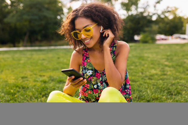 休闲年轻时尚微笑的黑人女子使用智能手机 通过无线耳机听音乐 在公园里玩得很开心女孩女人积极