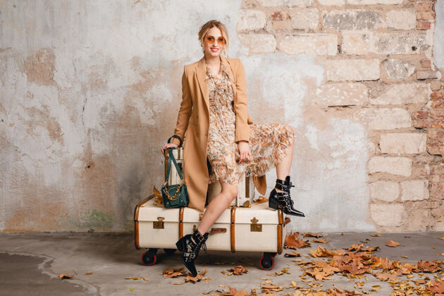 魅力在街上 穿着米色外套的金发美女坐在手提箱上对着墙年轻快乐时尚
