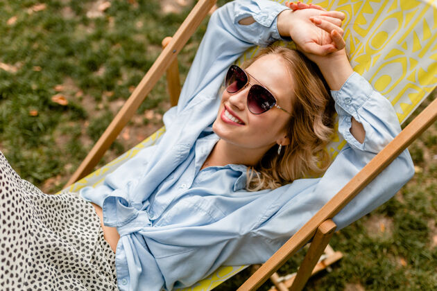年轻迷人的金发微笑的女人坐在躺椅上穿着夏天的衣服快乐穿太阳