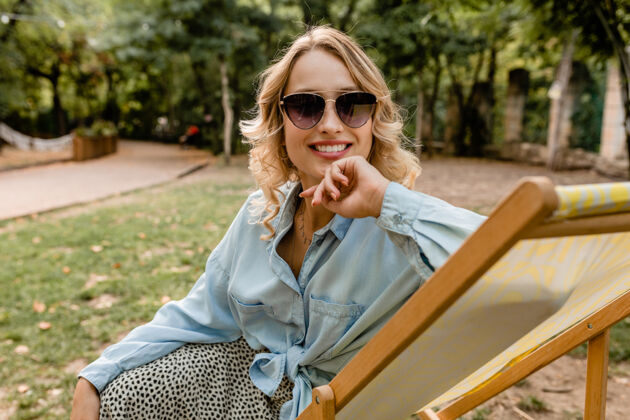 太阳镜迷人的金发微笑的女人 穿着时髦的服装 轻松地坐在躺椅上休息欢呼微笑