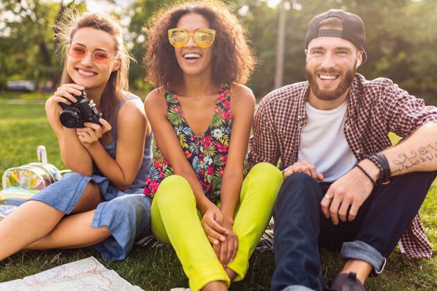 女孩快乐的年轻朋友们一起坐在公园里 男女一起玩 带着相机旅行 酷相机多种族