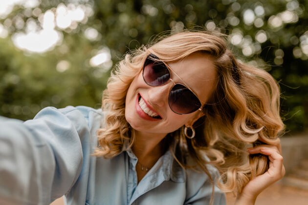 有趣迷人的金发微笑的白牙女人穿着夏装在公园里散步在电话里自拍模特肖像太阳镜