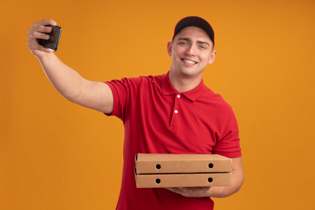 拿着微笑的年轻送货员穿着制服 戴着帽子 拿着披萨盒 在橙色的墙上自拍制服盒子帽子