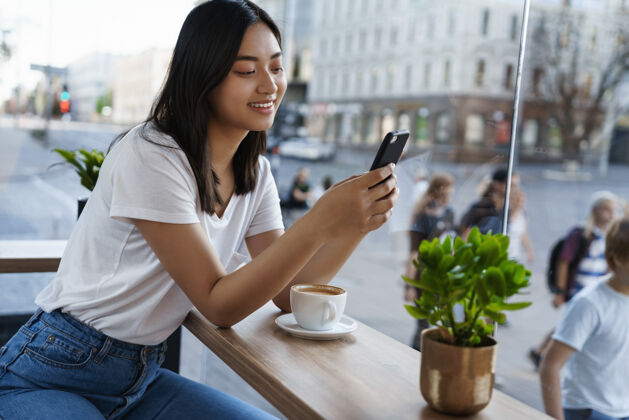 时尚美丽的亚洲女孩坐在窗前的城市咖啡馆里 一边用手机聊天 一边喝着咖啡 一边用智能手机微笑肖像喝酒