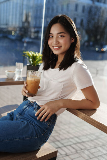 城市快乐的现代女孩坐在靠近窗户的咖啡馆里 倚在桌子上 喝着冰拿铁肖像休闲时尚