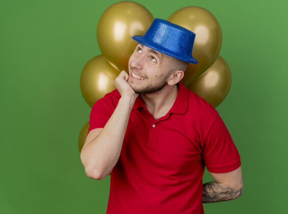派对面带微笑的年轻英俊的斯拉夫党人戴着党的帽子 背后拿着气球 手放在下巴上 在绿色的背景上看着孤立的复制空间下巴帽子背部