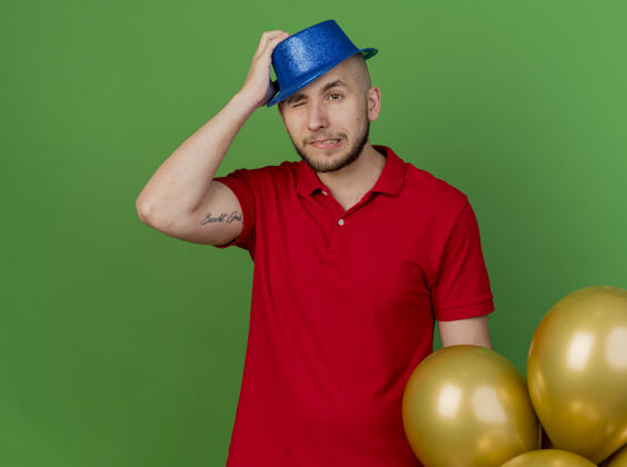 党遗憾的是年轻英俊的斯拉夫党人戴着党帽站在气球旁看着一边把手放在头上闭着一只眼睛隔离在绿色背景与复制空间气球家伙复制