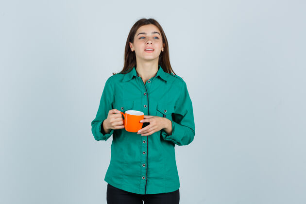持有年轻的女士拿着一杯橘色的茶 穿着衬衫 看上去很快乐 正对着窗外牙齿微笑女人