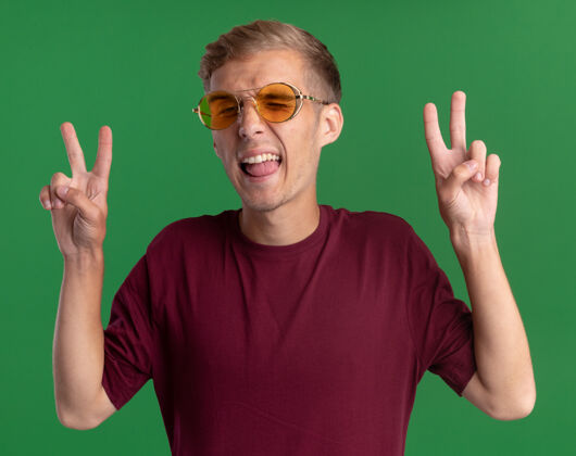 舌头快乐的年轻帅哥 穿着红衬衫 戴着眼镜 在绿色的墙上展示着舌头和和平的姿态年轻衬衫穿