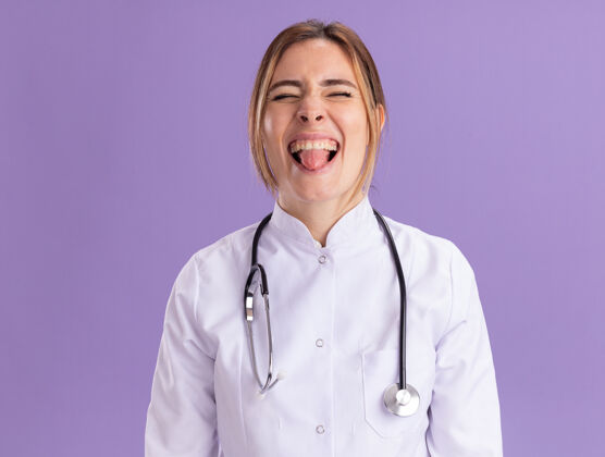 年轻年轻的女医生穿着医用长袍 带听诊器 舌头孤立在紫色的墙上女性欢乐医生