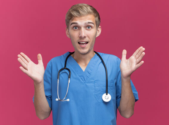 穿惊讶的年轻男医生穿着医生制服听诊器摊开双手隔离在粉红色的墙上年轻听诊器手
