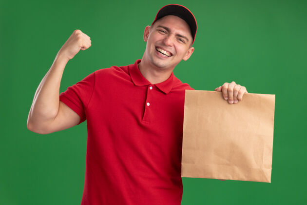 兴奋兴奋的年轻送货员穿着制服 戴着帽子 手里拿着一个纸食品包 在绿色的墙上显示“是”的手势帽子抱着男人